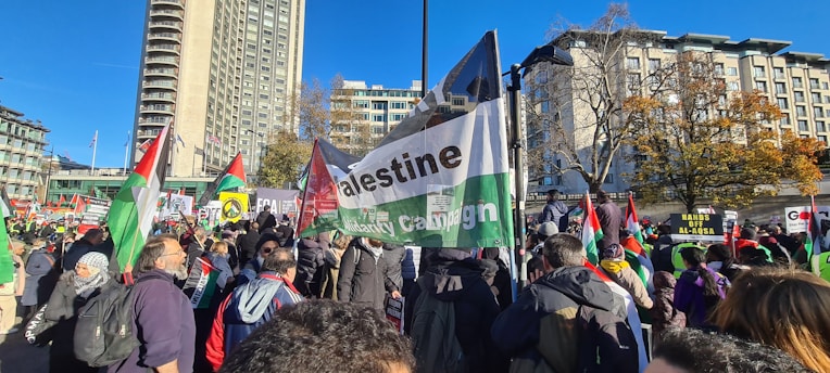 A large group of people gathered outdoors holding flags and signs. There are buildings and trees in the background, with a clear blue sky overhead. The signs and flags prominently feature the colors red, green, black, and white, and some have text related to a campaign or cause.