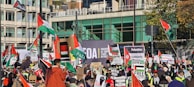 A large crowd of people gathered for a protest, holding various signs and flags related to Palestinian solidarity. Banners with messages such as 'Free Palestine' are prominent. The setting appears to be an urban area with modern buildings in the background.