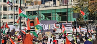 A large crowd of people gathered for a protest, holding various signs and flags related to Palestinian solidarity. Banners with messages such as 'Free Palestine' are prominent. The setting appears to be an urban area with modern buildings in the background.