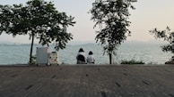 A tranquil scene by a waterfront, featuring two people sitting together on a paved area surrounded by trees. Their backs are turned toward the camera, and they are looking out over the water. There is a suitcase nearby, adorned with stickers. The distant skyline and mountains are visible under a clear sky.