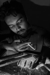 A person is sitting with eyes closed, holding a cigarette. The scene is dimly lit, creating a dramatic and introspective atmosphere. A table with several lines of a powdered substance and a card used for organizing the substance is visible. The shadows cast by the low light emphasize the contemplative mood.