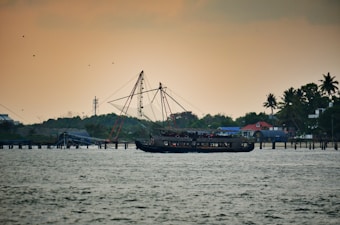 A traditional wooden fishing boat is sailing on water with an orange-tinted sky in the background. The boat is equipped with nets and rigging, and there are several birds flying above. The shoreline in the distance features lush greenery, a few small buildings with red and blue roofs, and scattered palm trees.
