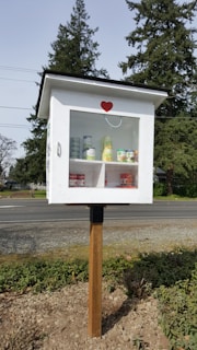 A small, white wooden box mounted on a pole, resembling a mini pantry or library. The box has a transparent door with a red heart decal on the front. Inside, there are various canned goods neatly arranged on two shelves. The background features tall evergreen trees and a road, suggesting an outdoor, community setting.