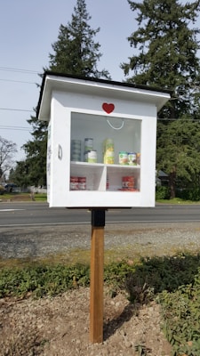 A small, white wooden box mounted on a pole, resembling a mini pantry or library. The box has a transparent door with a red heart decal on the front. Inside, there are various canned goods neatly arranged on two shelves. The background features tall evergreen trees and a road, suggesting an outdoor, community setting.