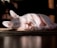 A hairless cat is lying on its side, basking in warm sunlight with its large, expressive eyes looking towards the camera. The cat's skin is pale, with visible folds and wrinkles. The light casts soft shadows on the floor and highlights the textures of the cat's skin.