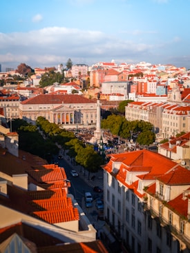 A vibrant cityscape featuring a central plaza with a tall monument surrounded by people and various trees. The buildings have distinctive terracotta roofs and colorful facades, creating a lively atmosphere. Streets are lined with cars, and the sky is partly cloudy with a hint of sunlight illuminating the scene.