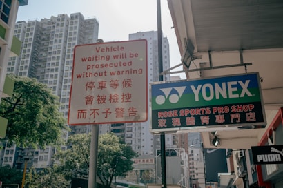 A busy urban street scene with tall residential buildings in the background. A traffic sign in both English and Chinese warns that vehicle waiting will be prosecuted. On the right, a storefront sign reads 'Yonex Rose Sports Pro Shop' with accompanying Chinese characters. Various brand names such as Puma are visible, suggesting a commercial area.