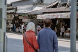 Two people with their backs turned are standing in front of a busy street market. They appear to be observing the market activities. The person on the left is wearing a red jacket, while the person on the right is wearing a blue shirt. Several people can be seen engaging in various activities at the market stalls, which are filled with products and signs. The market has a rustic and lived-in appearance.