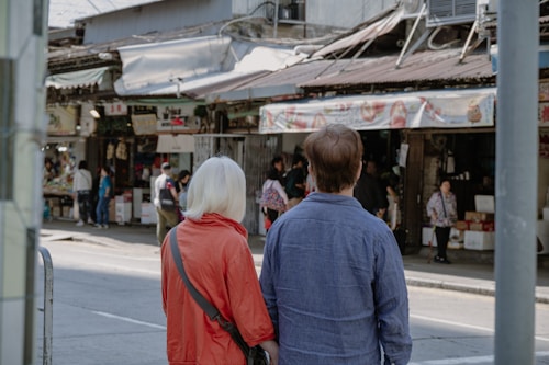 Two people with their backs turned are standing in front of a busy street market. They appear to be observing the market activities. The person on the left is wearing a red jacket, while the person on the right is wearing a blue shirt. Several people can be seen engaging in various activities at the market stalls, which are filled with products and signs. The market has a rustic and lived-in appearance.