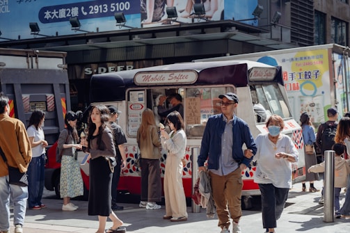 A bustling urban street scene with several people gathered around an ice cream truck labeled 'Mobile Softee'. The group includes individuals enjoying ice cream and engaging in casual conversation. The background features advertisements and building facades, indicating a lively city environment.
