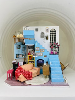 A detailed miniature model of a cozy living room featuring a blue staircase and a blue wall with various framed art pieces. There is a small orange checkered sofa with a red polka-dotted blanket draped over it, a fireplace with the word 'Fire' written on it, and a fluffy white rug on a wooden floor. Various small objects, including plants and pots, are arranged neatly, creating a homely atmosphere.