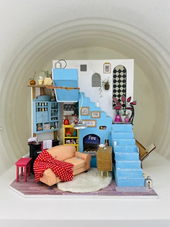 A detailed miniature model of a cozy living room featuring a blue staircase and a blue wall with various framed art pieces. There is a small orange checkered sofa with a red polka-dotted blanket draped over it, a fireplace with the word 'Fire' written on it, and a fluffy white rug on a wooden floor. Various small objects, including plants and pots, are arranged neatly, creating a homely atmosphere.