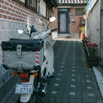 A white scooter is parked in a narrow alley with a brick wall on one side and a flower-lined sidewalk on the other. The scooter has a silver storage box mounted on the back and a windshield in front. The alley leads to a building with a dark door.