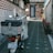 A white scooter is parked in a narrow alley with a brick wall on one side and a flower-lined sidewalk on the other. The scooter has a silver storage box mounted on the back and a windshield in front. The alley leads to a building with a dark door.