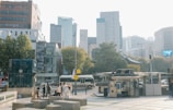 Urban scene featuring tall modern buildings in the background, a busy street with several vehicles, a group of people walking, and a small outdoor structure that appears to be a kiosk or bus stop. Trees with autumn foliage line the street, adding a touch of nature to the bustling city environment.