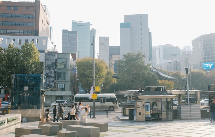 Urban scene featuring tall modern buildings in the background, a busy street with several vehicles, a group of people walking, and a small outdoor structure that appears to be a kiosk or bus stop. Trees with autumn foliage line the street, adding a touch of nature to the bustling city environment.