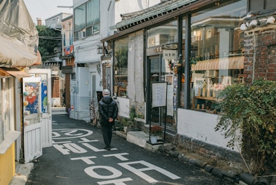 A narrow alleyway bordered by small shops and buildings with a person walking away, wearing a puffer jacket and beanie. The storefronts have large windows displaying various items, some signage in Korean, and plants add greenery to the scene. The atmosphere suggests a quaint, cozy neighborhood.
