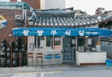 A small traditional Korean restaurant with a blue awning, displaying Korean text. The building has a tiled roof and is flanked by earthenware jars on the side. Three stools are placed outside for seating, creating a quaint street-side dining setup.