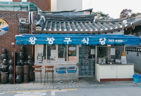 A small traditional Korean restaurant with a blue awning, displaying Korean text. The building has a tiled roof and is flanked by earthenware jars on the side. Three stools are placed outside for seating, creating a quaint street-side dining setup.
