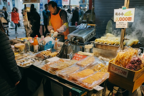A bustling street food stall with a variety of snacks on display, including packaged dried foods and skewered items. Steam rises from a grill where food is being cooked, and the vendor wears an orange apron. Shoppers and pedestrians are visible in the background, creating a lively market atmosphere.