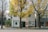A two-story building with a white facade is partially obscured by large trees with vibrant yellow leaves. The street is wide and asphalted, with a sidewalk lined with plants and a small fire hydrant. The building's signage indicates a business named 'NDIPRESS'. The windows are adorned with colorful abstract art pieces.