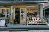 A quaint plant shop with a variety of potted plants and flowers displayed on shelves outside the entrance. The shop has a warm, inviting atmosphere with large glass windows showcasing more plants inside. The sign above reads 'Amélie' while the glass door has another sign that says 'howamy plant shop & cafe.' Several planters with green foliage are placed on the pavement, contributing to the shop's charming aesthetic.