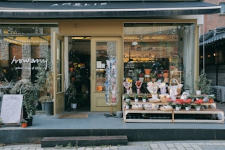 A quaint plant shop with a variety of potted plants and flowers displayed on shelves outside the entrance. The shop has a warm, inviting atmosphere with large glass windows showcasing more plants inside. The sign above reads 'Amélie' while the glass door has another sign that says 'howamy plant shop & cafe.' Several planters with green foliage are placed on the pavement, contributing to the shop's charming aesthetic.