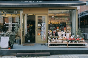 A quaint plant shop with a variety of potted plants and flowers displayed on shelves outside the entrance. The shop has a warm, inviting atmosphere with large glass windows showcasing more plants inside. The sign above reads 'Amélie' while the glass door has another sign that says 'howamy plant shop & cafe.' Several planters with green foliage are placed on the pavement, contributing to the shop's charming aesthetic.