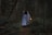 A person dressed in a flowing, light-colored cloak walks through a dimly lit forest while holding a glowing lantern. The dense forest is filled with dark green foliage and tall, slender trees. The atmosphere is mysterious and somewhat magical, as the lantern casts a warm, soft light amidst the dark surroundings.