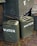 Several army green jerry cans labeled 'WATER' are stacked alongside wooden crates. The crates display text reading 'CORPS OF ENGINEERS' and 'U.S. ARMY SUPPLIES'. The scene is set on a brick pavement with some moss visible between the bricks.