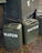 Several army green jerry cans labeled 'WATER' are stacked alongside wooden crates. The crates display text reading 'CORPS OF ENGINEERS' and 'U.S. ARMY SUPPLIES'. The scene is set on a brick pavement with some moss visible between the bricks.