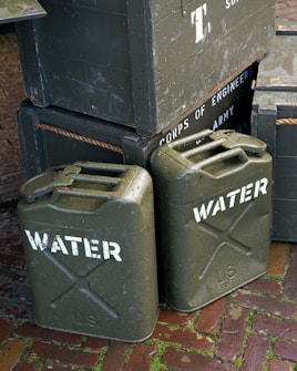 Several army green jerry cans labeled 'WATER' are stacked alongside wooden crates. The crates display text reading 'CORPS OF ENGINEERS' and 'U.S. ARMY SUPPLIES'. The scene is set on a brick pavement with some moss visible between the bricks.