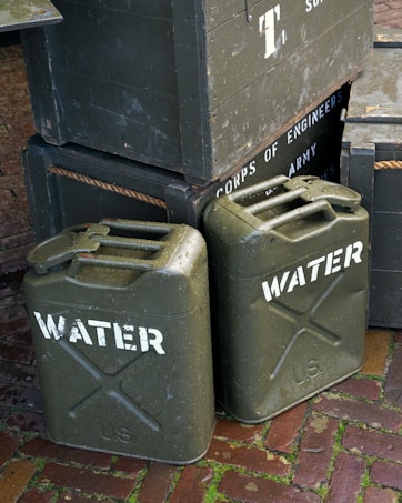 Several army green jerry cans labeled 'WATER' are stacked alongside wooden crates. The crates display text reading 'CORPS OF ENGINEERS' and 'U.S. ARMY SUPPLIES'. The scene is set on a brick pavement with some moss visible between the bricks.