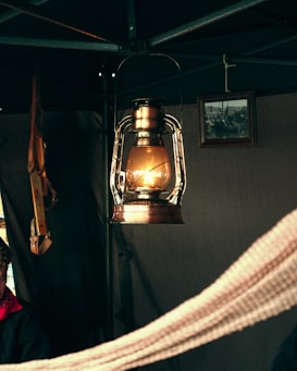 A vintage metal lantern emits a warm glow, casting light within a tent-like setting. A framed black-and-white photograph hangs in the background, adding a nostalgic touch. A piece of fabric is draped across the foreground, with some leather straps visible on the side.