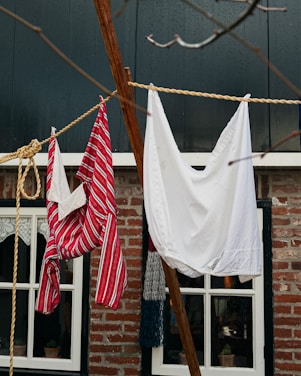 Laundry hangs on a clothesline in front of a rustic brick house. One item is a red and white striped garment, while the other is a plain white cloth. The background features windows with lace curtains, framed in black.