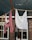 Laundry hangs on a clothesline in front of a rustic brick house. One item is a red and white striped garment, while the other is a plain white cloth. The background features windows with lace curtains, framed in black.