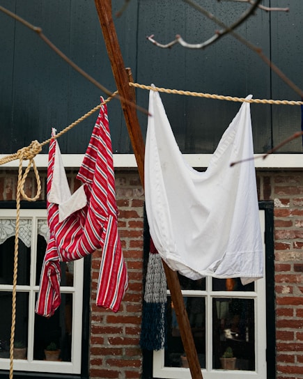 Laundry hangs on a clothesline in front of a rustic brick house. One item is a red and white striped garment, while the other is a plain white cloth. The background features windows with lace curtains, framed in black.