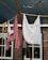 Laundry hangs on a clothesline in front of a rustic brick house. One item is a red and white striped garment, while the other is a plain white cloth. The background features windows with lace curtains, framed in black.