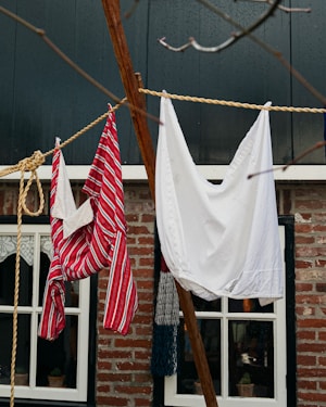 Laundry hangs on a clothesline in front of a rustic brick house. One item is a red and white striped garment, while the other is a plain white cloth. The background features windows with lace curtains, framed in black.