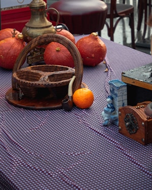 A collection of rustic and vintage items arranged on a checkered tablecloth. There are several orange pumpkins, a brass teapot with a handle, a wooden basket, a small orange, a wooden pipe, and a decorative ceramic figure. The scene is set outdoors with wooden chairs in the background.