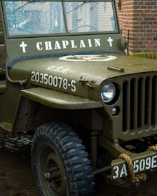 A vintage military jeep with the word 'CHAPLAIN' painted on the front, displaying a military serial number and insignia. Raindrops are present on the windshield, indicating recent rain. The jeep is parked in a brick-paved area with a rope tied around the front bumper.