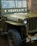 A vintage military jeep with the word 'CHAPLAIN' painted on the front, displaying a military serial number and insignia. Raindrops are present on the windshield, indicating recent rain. The jeep is parked in a brick-paved area with a rope tied around the front bumper.