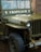 A vintage military jeep with the word 'CHAPLAIN' painted on the front, displaying a military serial number and insignia. Raindrops are present on the windshield, indicating recent rain. The jeep is parked in a brick-paved area with a rope tied around the front bumper.