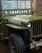 A vintage military jeep with the word 'CHAPLAIN' painted on the front, displaying a military serial number and insignia. Raindrops are present on the windshield, indicating recent rain. The jeep is parked in a brick-paved area with a rope tied around the front bumper.