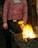 A person wearing a striped red and purple shirt engaged in blacksmithing work. They are standing beside a worktable with glowing hot coals and flames. A large hammer is resting on the table, and the individual appears focused on their task.