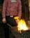 A person wearing a striped red and purple shirt engaged in blacksmithing work. They are standing beside a worktable with glowing hot coals and flames. A large hammer is resting on the table, and the individual appears focused on their task.