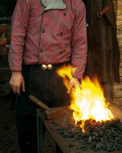 A person wearing a striped red and purple shirt engaged in blacksmithing work. They are standing beside a worktable with glowing hot coals and flames. A large hammer is resting on the table, and the individual appears focused on their task.