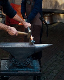 A person is using a hammer to forge a piece of glowing metal on an anvil. The setting appears to be an outdoor workshop with a cobblestone ground and backdrop materials like aprons and tools. The hands are the primary focus, emphasizing the craftsmanship involved in blacksmithing.