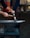A person is using a hammer to forge a piece of glowing metal on an anvil. The setting appears to be an outdoor workshop with a cobblestone ground and backdrop materials like aprons and tools. The hands are the primary focus, emphasizing the craftsmanship involved in blacksmithing.
