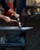A person is using a hammer to forge a piece of glowing metal on an anvil. The setting appears to be an outdoor workshop with a cobblestone ground and backdrop materials like aprons and tools. The hands are the primary focus, emphasizing the craftsmanship involved in blacksmithing.